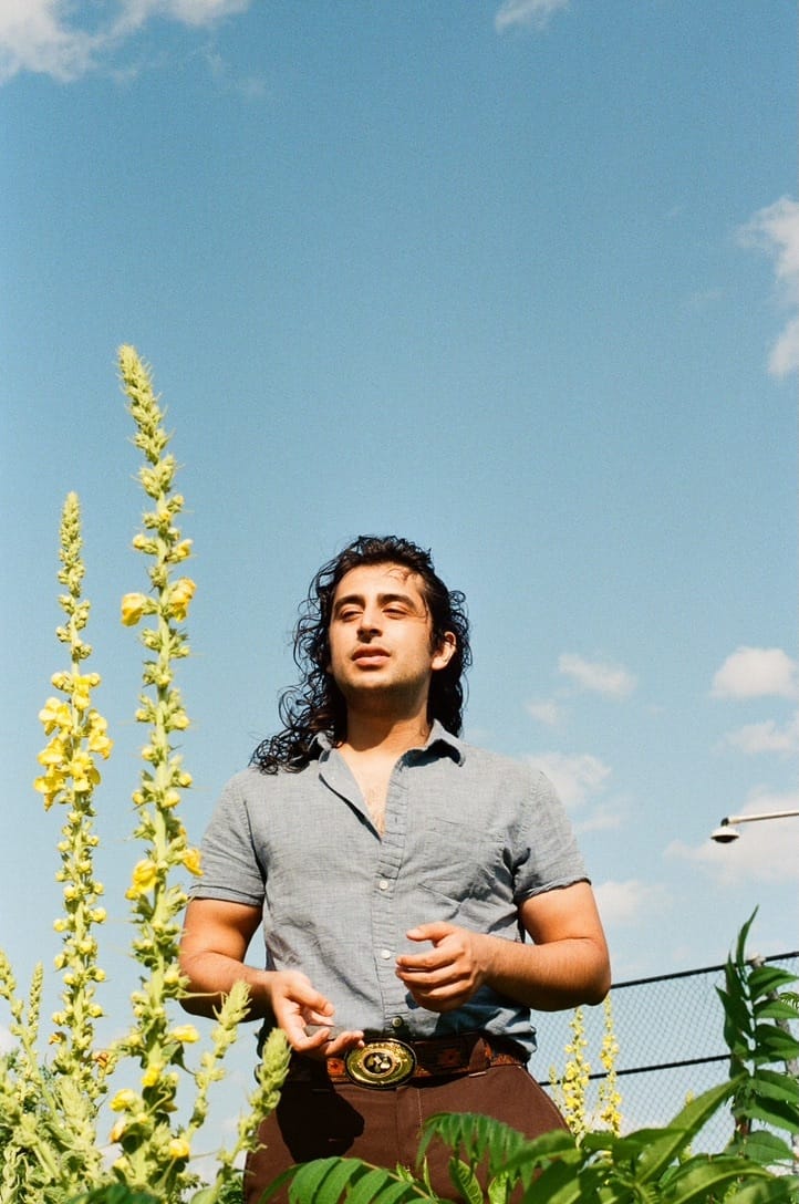 Andres Cordoba, a Colombian-Peruvian man with shoulder-length wavy, black hair stands in a blue-collared shirt with his hands clasped in front of him. Looking towards the sun, his eyes are closed and he is smiling. The photo is from a lower-angle and behind him is a slightly-clouded blue sky. In the foreground, covering his lower-half, there is foliage.
