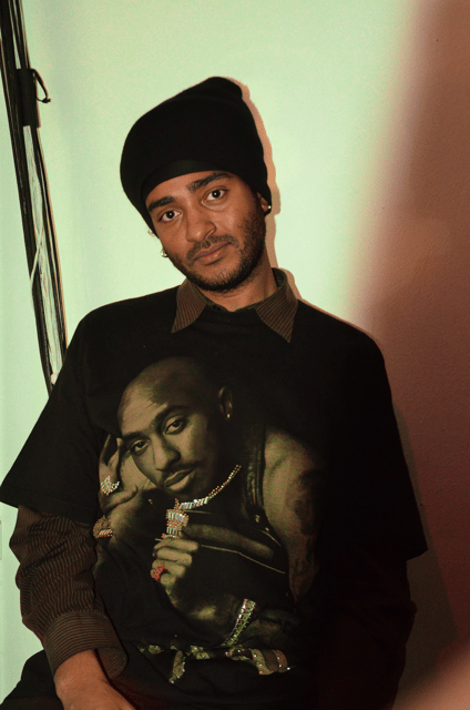 Jared Brown, a Black person wearing a black beanie, a 2pac shirt over a brown button up sits against a wall