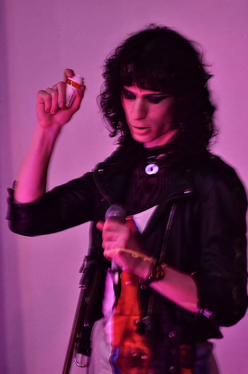 Noa Micaela Fields, a white trans woman saturated in magenta light, shakes a bottle of estrogen pills in a performance at Elastic Arts.