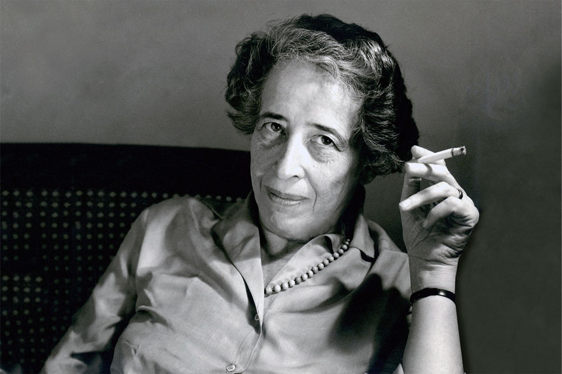 A black-and-white photograph of Hannah Arendt facing the camera while holding a lit cigarette.